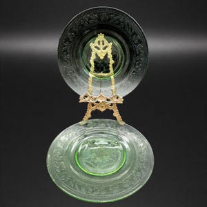 May include: Two green glass plates with floral patterns are displayed on a gold-colored metal stand. The plates have a decorative rim and a central design. The stand is ornate, with a detailed design. The background is black.