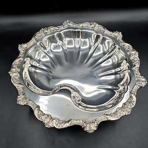 Puede incluir: Una fuente plateada ornamentada con un diseño en forma de concha. La fuente tiene un borde festoneado con detalles de volutas y una depresión central curvada. La superficie reflectante sugiere un acabado metálico pulido, adecuado para servir alimentos o como pieza decorativa.
