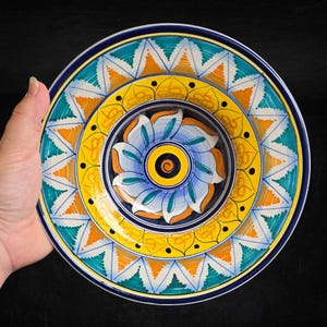 May include: A round ceramic plate featuring a hand-painted design with a central blue, white, and orange floral motif. The border displays concentric rings of yellow, teal, and orange geometric patterns, including triangles and stylized petals.