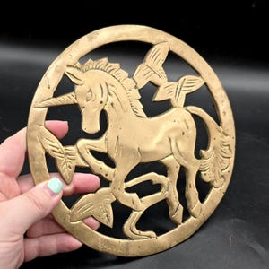 May include: A brass-colored, circular trivet featuring a unicorn design. The unicorn is surrounded by a decorative border with leaves and butterflies. The trivet is held by a hand against a black background.