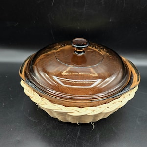 May include: A brown glass casserole dish with a matching lid, resting in a woven basket. The dish has a rounded shape and a dark amber hue. The basket is made of light-colored woven material.