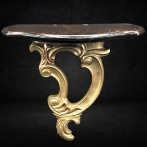 May include: A wall-mounted console shelf featuring a dark brown, semi-circular wooden top surface supported by a single, ornate gold-colored bracket with curved, scroll-like detailing extending downward from the center of the shelf.