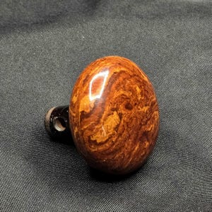 May include: A brown and orange, oval-shaped bottle stopper with a swirling wood grain pattern. The stopper has a dark brown base with a small hole. The product is set against a dark gray background.