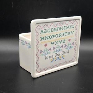 Può includere: Una scatola di ceramica bianca con un disegno a punto croce. Il disegno include l'alfabeto, cuori e la frase "Bless This Child" in lettere nere. La scatola ha una forma quadrata con un fronte rialzato.