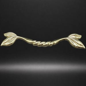 May include: A gold-colored, decorative cabinet pull with a mermaid tail design. The pull features two stylized mermaid tail ends connected by a twisted, rope-like center. The hardware is set against a dark background.