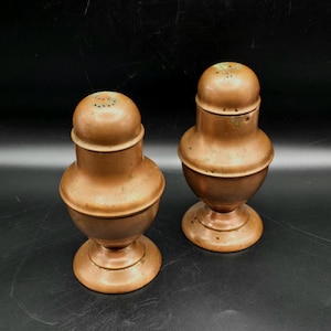 May include: Two antique copper salt and pepper shakers. Each shaker has a bulbous body, a pedestal base, and a perforated top. The copper has a warm, reddish-brown hue with some areas of patina. The shakers are displayed against a dark background.
