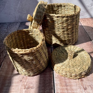 May include: Two woven baskets made of natural straw with a lid. The baskets are round and have a natural, light brown color.