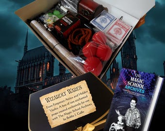 May include: An open box filled with magic props, including playing cards, red sponge balls, a small wooden chest, and a deck of cards. A book titled "The Magic School Archives" is also visible, along with a black envelope with the text "Wizardly Wares".