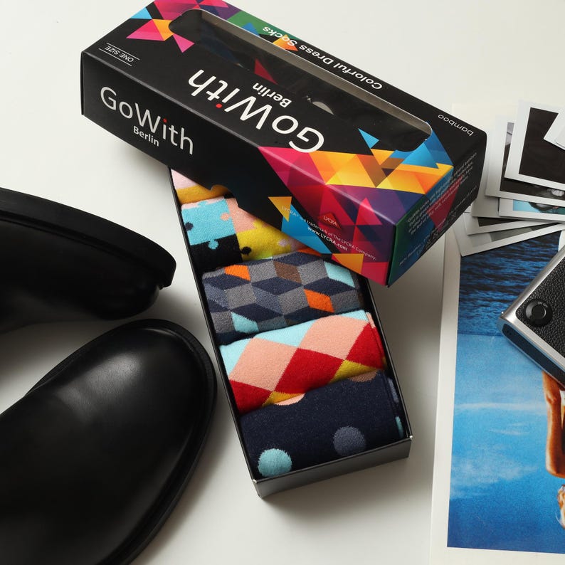 May include: A box set of colorful dress socks from Go With Berlin, featuring various geometric and polka dot patterns. The box is black with a colorful geometric design and the words "Go With Berlin" printed on it. Black leather shoes are in the foreground.