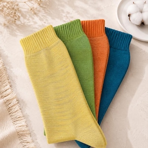 May include: A set of four crew socks featuring a ribbed cuff design. The socks are presented in a vibrant color palette consisting of solid yellow, bright green, burnt orange, and deep teal, each showcasing a fine knit texture throughout the fabric.