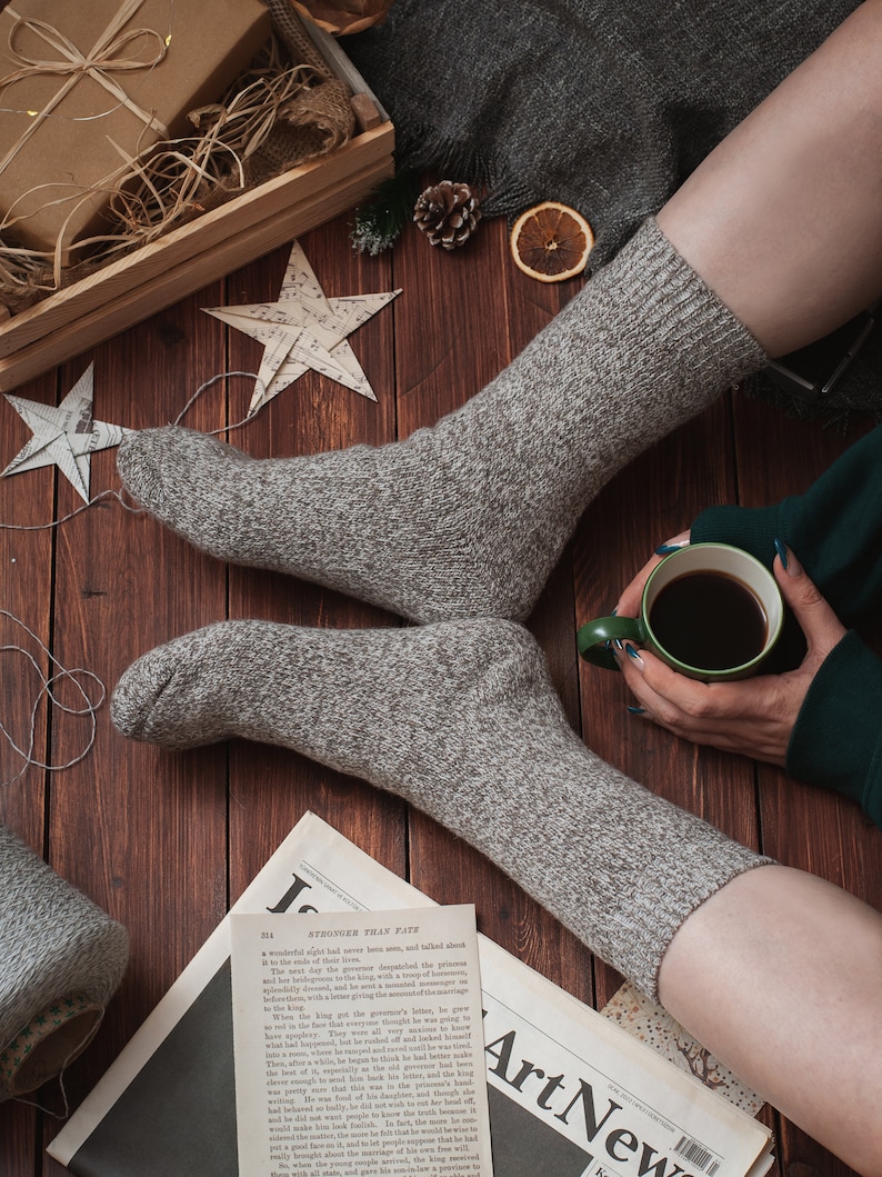 May include: A pair of grey knitted socks with a cup of coffee and a newspaper on a wooden surface. The newspaper has the headline "STRONGER THAN FATE".