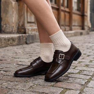 K&ouml;nnte beinhalten: Ein Paar dunkelbraune Leder-Loafer mit Schnallenverschluss. Die Schuhe haben eine eckige Zehenpartie und eine dicke schwarze Sohle. Die Person tr&auml;gt beige Socken.