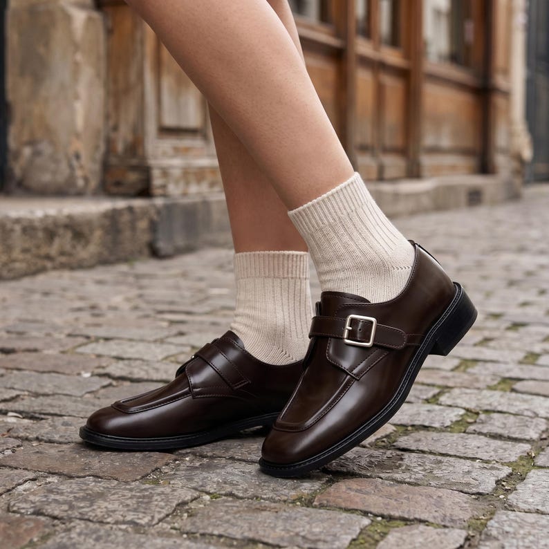 K&ouml;nnte beinhalten: Ein Paar dunkelbraune Leder-Loafer mit Schnallenverschluss. Die Schuhe haben eine eckige Zehenpartie und eine dicke schwarze Sohle. Die Person tr&auml;gt beige Socken.