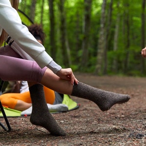 May include: A woman wearing brown socks sits on a green camping chair in a woodland setting. The socks are made of a soft, textured material and have a comfortable fit.