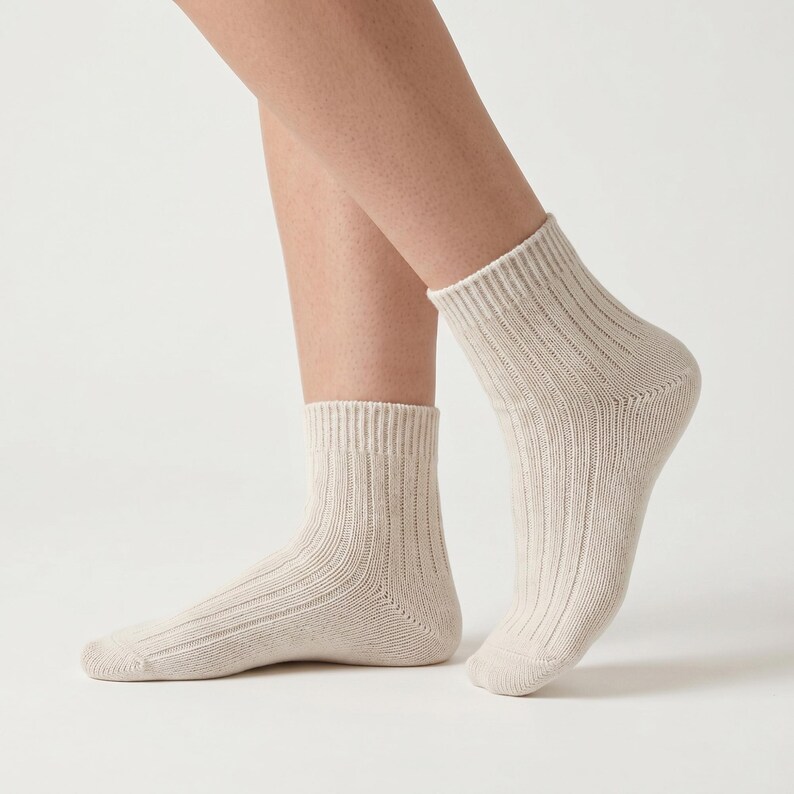 K&ouml;nnte beinhalten: Ein Paar beige gerippte Kn&ouml;chelsocken. Die Socken bestehen aus einem weichen Material und haben ein geripptes Design. Die Socken werden an den F&uuml;&szlig;en einer Person getragen, wobei die Beine im Hintergrund sichtbar sind. Die Socken sind neutral gef&auml;rbt.
