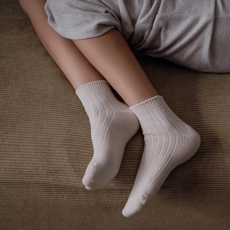K&ouml;nnte beinhalten: Ein Paar cremefarbene gerippte Socken. Die Socken bestehen aus einem weichen Material und haben ein geripptes Design. Die Socken werden an den F&uuml;&szlig;en einer Person getragen, wobei die Beine und ein Teil des K&ouml;rpers sichtbar sind. Der Hintergrund ist eine braune Oberfl&auml;che.