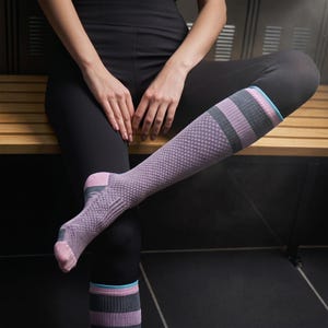 May include: A person wearing black leggings and a lavender knee-high sock with a checkered pattern. The sock has gray and pink stripes at the top, with a light blue accent. The person is sitting on a wooden bench.