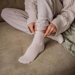 K&ouml;nnte beinhalten: Nahaufnahme von gerippten, cremefarbenen Kn&ouml;chelsocken. Die Socken werden von einer Person angepasst, die einen hellgrauen Pullover und eine passende Hose tr&auml;gt. Die Szene spielt sich auf einer strukturierten, neutral get&ouml;nten Oberfl&auml;che ab.