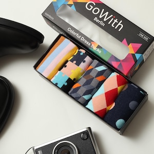 May include: A box set of colorful dress socks with various patterns, including stripes, geometric shapes, and polka dots. The box is black with the text "Go With Berlin" and "Colorful Dress Socks". A black shoe and a camera are also in the image.