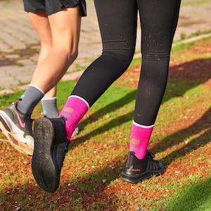 May include: Two people wearing black leggings and colorful socks while running on a red and green surface. The person in the foreground is wearing pink socks with white and orange accents. The person in the background is wearing gray socks with blue and white accents.