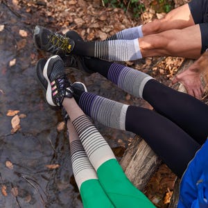 May include: Three pairs of legs wearing colorful striped socks and leggings. The socks are black, white, and gray with purple stripes. The leggings are black with purple and gray stripes. The socks and leggings are worn with black shoes.