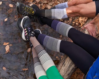 May include: Three pairs of legs wearing colorful striped socks and leggings. The socks are black, white, and gray with purple stripes. The leggings are black with purple and gray stripes. The socks and leggings are worn with black shoes.
