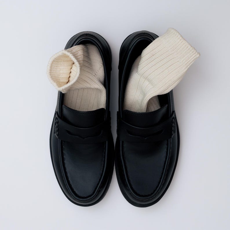 K&ouml;nnte beinhalten: Ein Paar schwarze Leder-Loafer mit cremefarbenen Socken. Die Schuhe haben ein klassisches Design mit runder Spitze und einem Riemen &uuml;ber dem Spann. Die Socken sind gerippt und ragen &uuml;ber den Schuhschaft hinaus. Draufsicht.