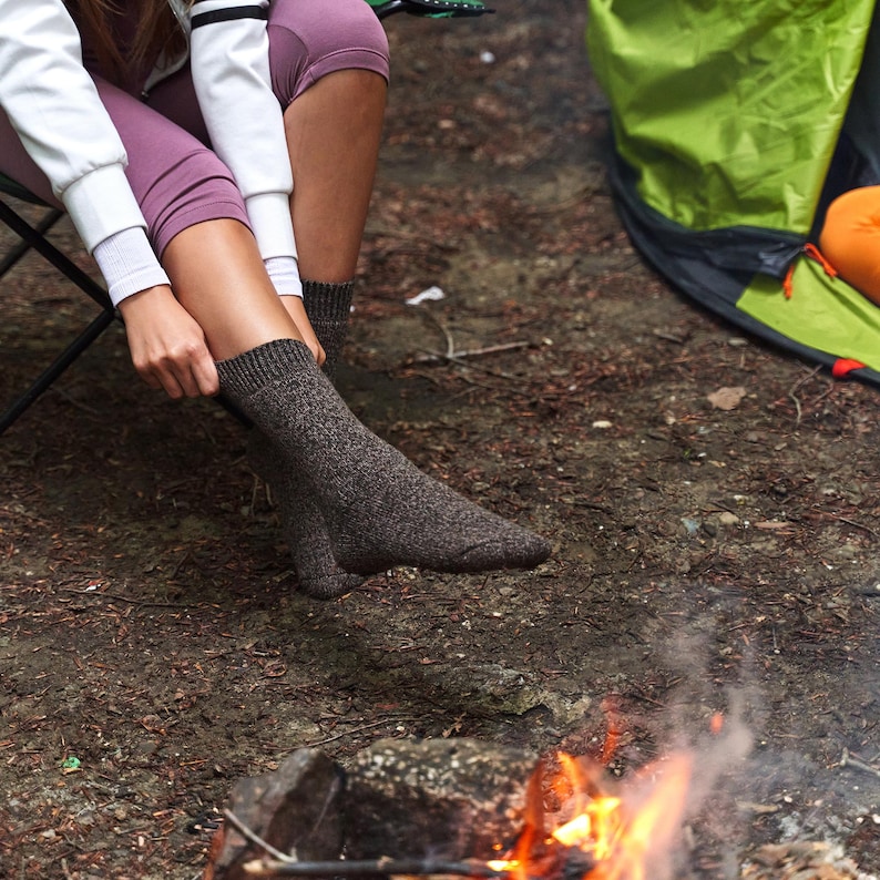 May include: A person wearing brown socks sits by a campfire in a woodland setting. The person is wearing a white jacket and purple trousers. The fire is small and surrounded by rocks.