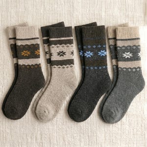 May include: A collection of five pairs of wool socks in various shades of gray, brown, and black. Each pair features a patterned design with geometric shapes and contrasting colors. The socks are arranged on a white, textured surface.