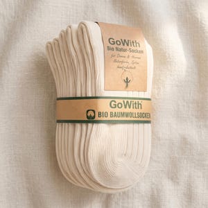 K&ouml;nnte beinhalten: Ein B&uuml;ndel cremefarbener gerippter Socken, umwickelt mit einem braunen Papierband. Das Band und das angebrachte Etikett zeigen den Text "GoWith" und deutschen Text. Die Socken sind aus Bio-Baumwolle.