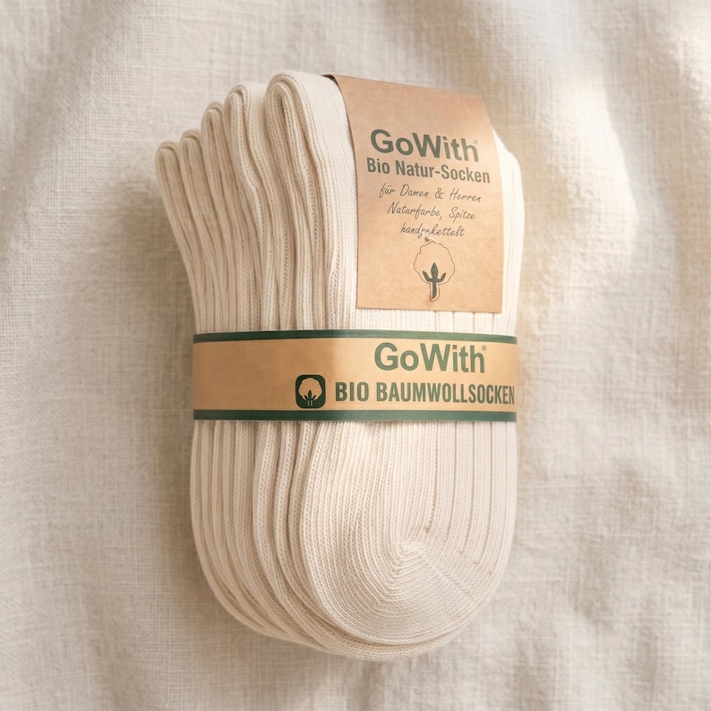 K&ouml;nnte beinhalten: Ein B&uuml;ndel cremefarbener gerippter Socken, umwickelt mit einem braunen Papierband. Das Band und das angebrachte Etikett zeigen den Text "GoWith" und deutschen Text. Die Socken sind aus Bio-Baumwolle.