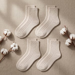 K&ouml;nnte beinhalten: Vier Paar cremefarbene gerippte Socken sind auf einer neutral get&ouml;nten Oberfl&auml;che angeordnet. Die Socken sind gefaltet und paarweise ausgestellt. Baumwollzweige mit wei&szlig;en Bl&uuml;ten sind um die Socken herum platziert und f&uuml;gen der Komposition ein nat&uuml;rliches Element hinzu.