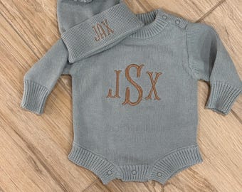 May include: A light blue knitted baby outfit set, including a romper and a beanie hat with a pom-pom. The romper features long sleeves and snap closures, with the initials "JSX" embroidered in a contrasting brown color. The beanie is embroidered with the name "JAX".