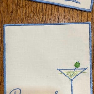 K&ouml;nnte beinhalten: Quadratische wei&szlig;e Cocktailservietten mit hellblauem Stickrand. Jede Serviette zeigt ein Martiniglas mit einer Olive und dem Namen "Romicks" in hellblauer Schrift gestickt. Die Servietten liegen auf einer Holzoberfl&auml;che.