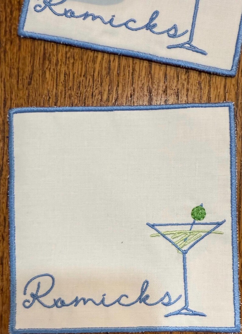 K&ouml;nnte beinhalten: Quadratische wei&szlig;e Cocktailservietten mit hellblauem Stickrand. Jede Serviette zeigt ein Martiniglas mit einer Olive und dem Namen "Romicks" in hellblauer Schrift gestickt. Die Servietten liegen auf einer Holzoberfl&auml;che.