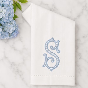 May include: A white folded hand towel featuring a light blue embroidered monogram letter S with a striped pattern. The towel has a subtle textured weave and a horizontal row of decorative hemstitch detailing near the bottom edge.