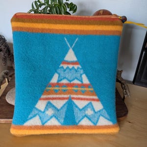 May include: A turquoise zippered pouch with a Native American teepee design. The pouch features a white teepee with orange and blue accents. The top and bottom edges are orange, and the zipper pull is yellow.