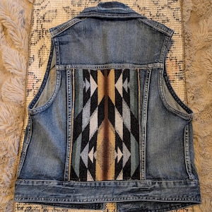 May include: A denim vest with a colorful geometric pattern on the back. The vest is made of blue denim and features a woven fabric panel with a design in shades of brown, black, gray, and teal. The vest has a classic collar and button closure.