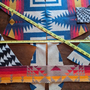 May include: Various pieces of fabric with geometric patterns in blue, white, red, yellow, and orange are laid out on a wooden surface. A yellow measuring tape shows measurements in inches. The fabric pieces appear to be cut for a garment.