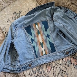May include: Light blue denim jacket with a colorful geometric design on the back. The design features shades of blue, teal, orange, and cream. The jacket has button closures and pockets.