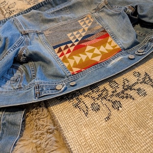 May include: A light blue denim jacket with a colorful geometric patch on the back. The patch features triangles in shades of red, yellow, and brown, with blue and white accents. The jacket has silver buttons and classic denim details.
