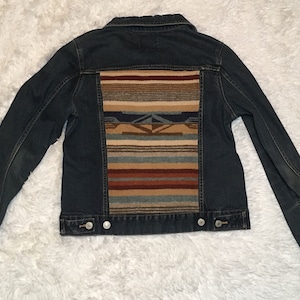 May include: A dark blue denim jacket with a colorful woven pattern on the back. The pattern is a traditional Native American design with stripes and geometric shapes in shades of brown, red, blue, and beige.