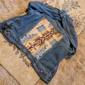 May include: A denim jacket with a colorful geometric pattern on the back. The jacket is a medium wash blue with metal buttons. The geometric pattern features shades of brown, blue, and cream.