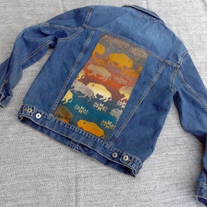 May include: A blue denim jacket with a colorful patterned fabric patch on the back. The patch features a repeating design of stylized buffalo and geometric shapes in shades of orange, yellow, blue, and green.