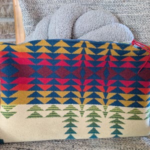 May include: A rectangular, colorful patterned pillow with a geometric design. The pillow features a cream-colored base with green tree-like shapes, and a vibrant upper section with blue, red, yellow, and orange triangles and diamonds. A red string is attached to the top corner.