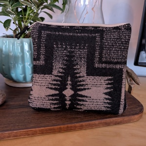 May include: A small, rectangular pouch with a black and tan geometric pattern. The pouch has a beige zipper and a matching beige lining. The pouch is made of a woven fabric and sits on a wooden tray.