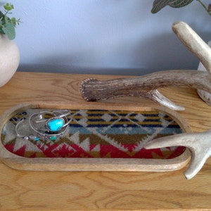 May include: A wooden tray with a patterned fabric lining, featuring a turquoise stone ring and other jewelry. The tray is next to a white antler on a wooden surface.