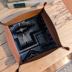 May include: A square, brown leather catchall tray with a patterned fabric interior in shades of black, gray, and white. The tray has curved edges and is sitting on a wooden surface. Books are visible in the background.