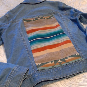 May include: A light blue denim jacket with a colorful woven patch on the back. The patch features a geometric pattern in shades of red, orange, blue, green, and beige.