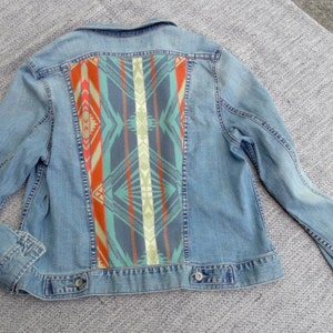 May include: A light blue denim jacket with a colorful geometric pattern on the back. The pattern features shades of red, orange, green, blue, and white.